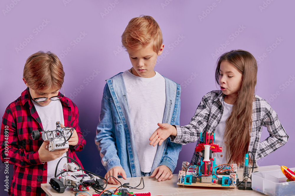 Fototapeta premium portrait of enthusiastic three kids working as team, assembling robots. three future engineers look at table full of robotics, two boys and one girl