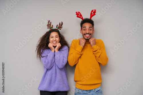 Overjoyed lovely young dark skinned curly couple looking happily at camera with wide smile and keeping raised fists under their chins, wearing holiday hoops and woolen sweaters over grey background
