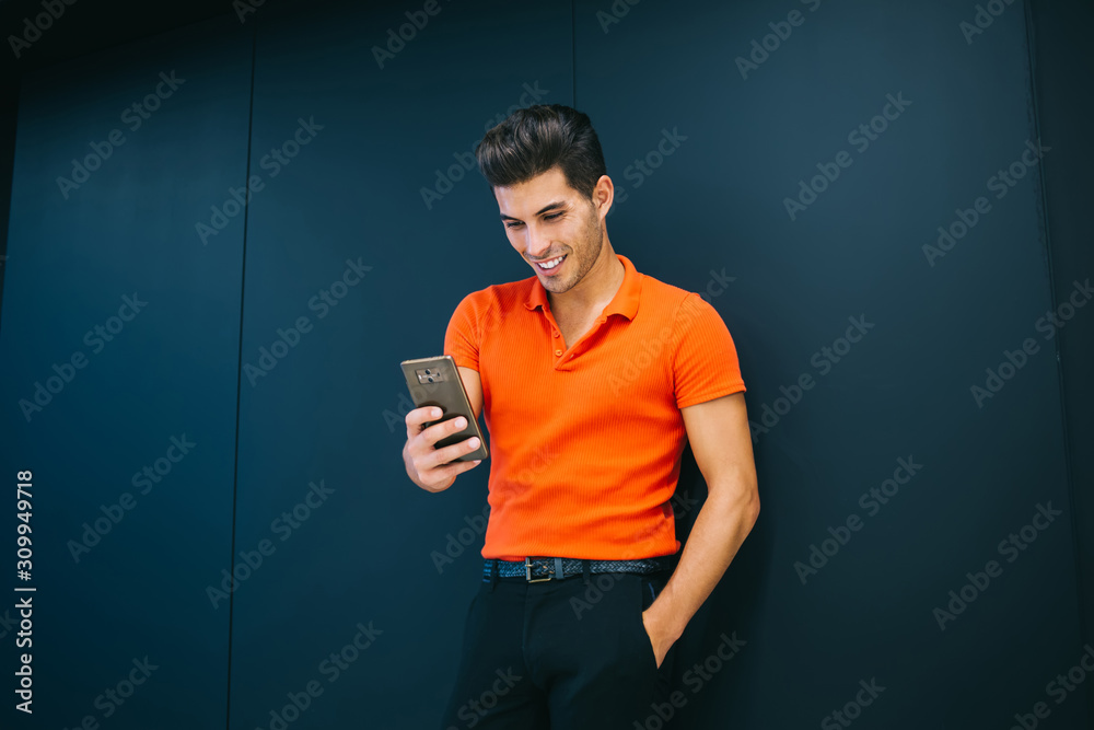 Smiling caucasian hipster guy in stylish wear watching video on smartphone during free time on promotional background, happy successful man in trendy outfit using mobile phone for communication Smiling caucasian hipster guy in stylish wear watching video on smartphone during free time on promotional background, happy successful man in trendy outfit using mobile phone for communication