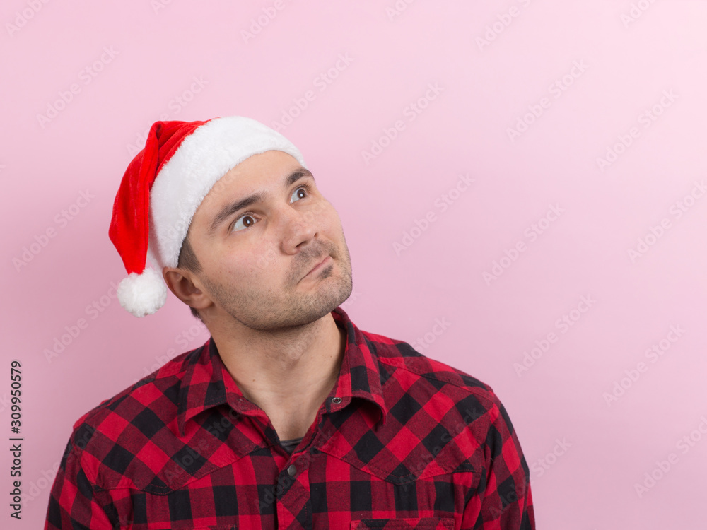 Emotions on the face, pensive, reflection, plan, idea. A man in a plaid rabbit and a Christmas red hat, on a pink background, copy space. Emotions on the face, pensive, reflection, plan, idea. A man in a plaid rabbit and a Christmas red hat, on a pink background, copy space.