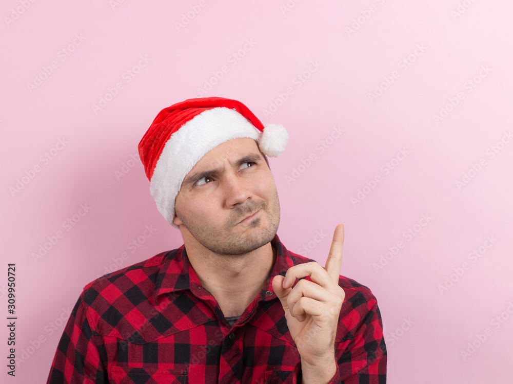 Emotions on the face, angry, bad, bandit, plotting evil. A man in a plaid rabbit and a Christmas red hat, on a pink background, copy space. Emotions on the face, angry, bad, bandit, plotting evil. A man in a plaid rabbit and a Christmas red hat, on a pink background, copy space.