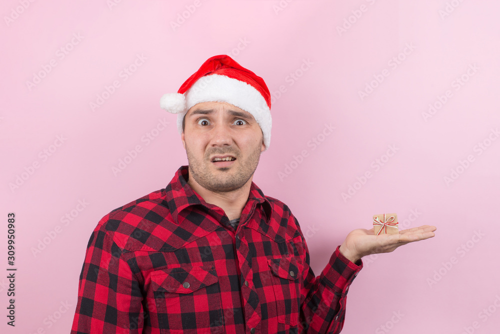 An unhappy, upset man in a red hat, holds a small gift with disgust on his face. Negative human emotions at christmas. Pink background, copy space. An unhappy, upset man in a red hat, holds a small gift with disgust on his face. Negative human emotions at christmas. Pink background, copy space.