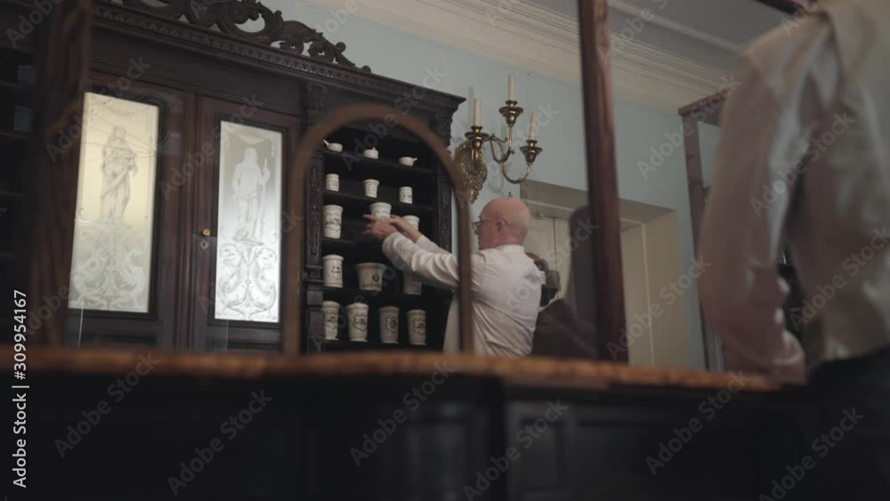 Senior Caucasian pharmacist taking old-fashioned container with medicine from shelf and talking to customer. 19th century drugstore, retro, vintage, pharmaceutical museum. Senior Caucasian pharmacist taking old-fashioned container with medicine from shelf and talking to customer. 19th century drugstore, retro, vintage, pharmaceutical museum.