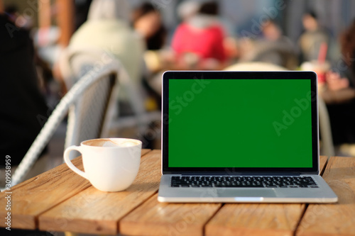 close up one green screen laptop computer on wooden table with a cup of coffee. blur sidewalk cafe and people background