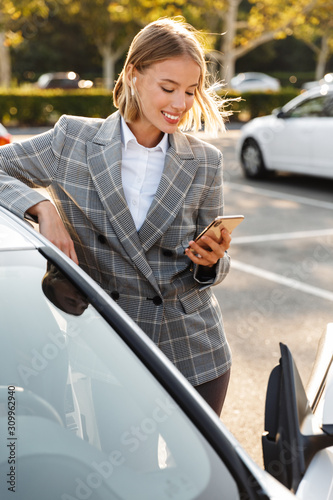 Fotografie Photo of businesswoman using earpod and cellphone while leaning on car
