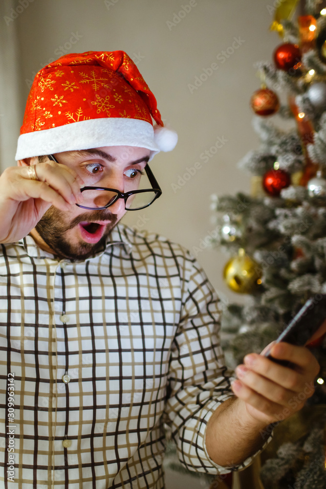 Portrait of a shocked man holding the phone in his hand, near a Christmas tree. Portrait of a shocked man holding the phone in his hand, near a Christmas tree.