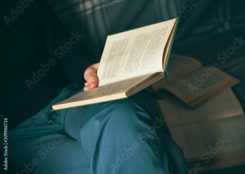 A man in blue pants and a black hoodie sits on a couch with open books scattered around, reading an encyclopedia.