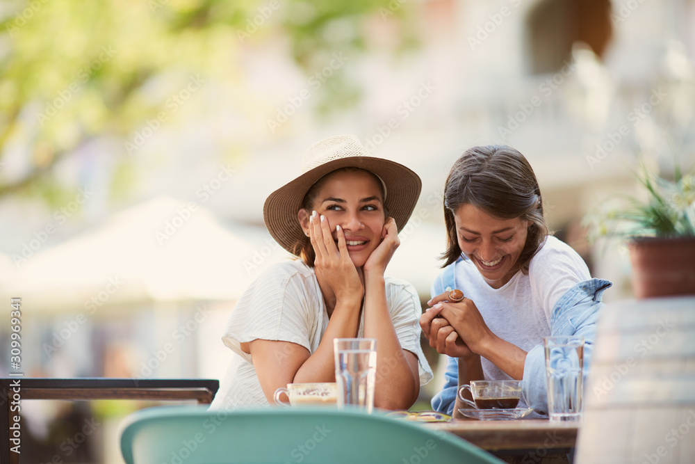  Two beautiful caucasian girls sitting in cafe outside and gossiping. On table are cups of coffee and grasses with water. Summer time.