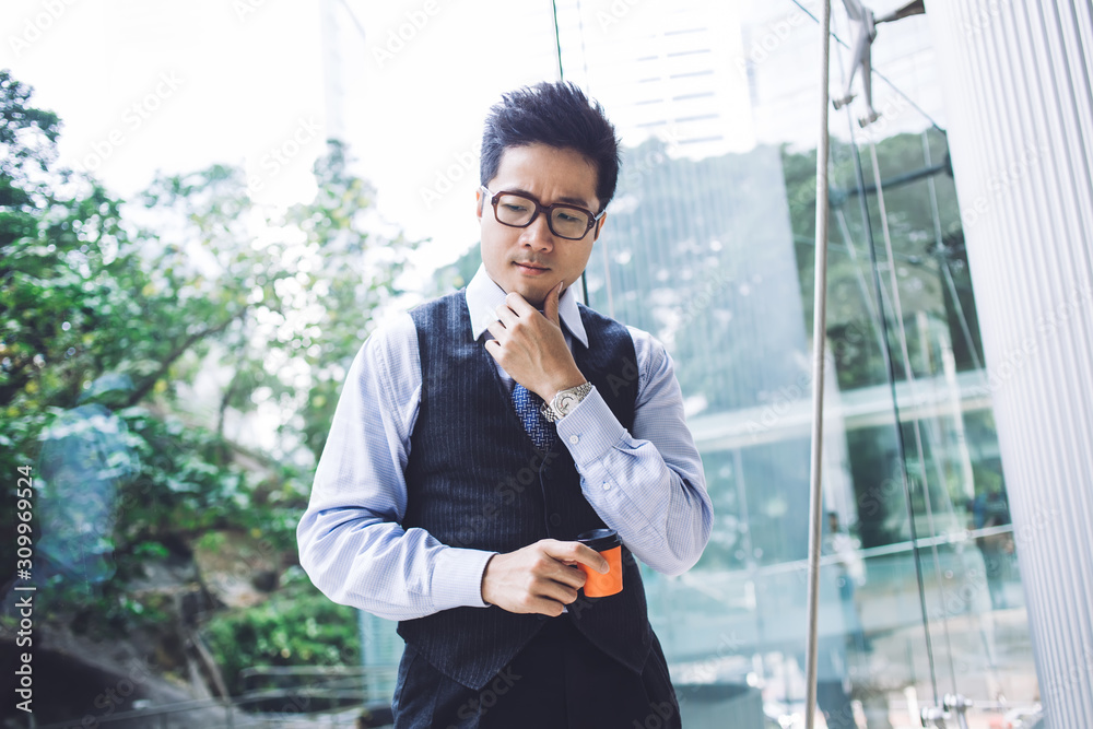 Thoughtful Asian businessman drinking coffee in disposable cup in building lobby Thoughtful Asian businessman drinking coffee in disposable cup in building lobby