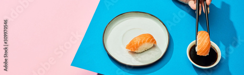 cropped view of woman putting fresh nigiri with salmon into soy sauce on blue, pink background, panoramic shot