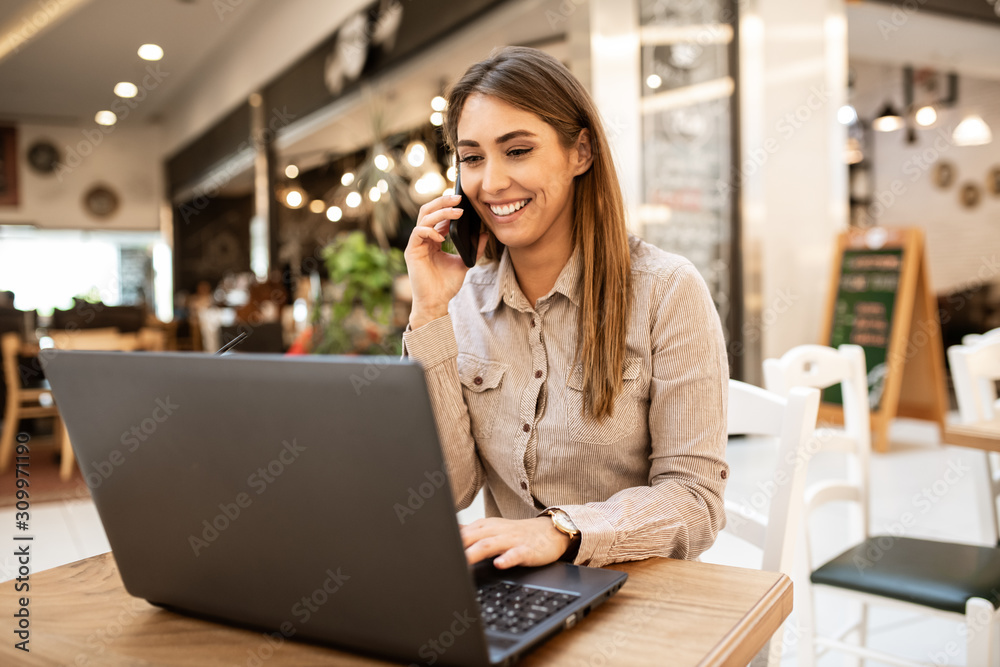 Gorgeous young caucasian woman with brown hair smiling and talking on her smart phone while browsing on her laptop. She is relaxing and sitting in a coffee shop.