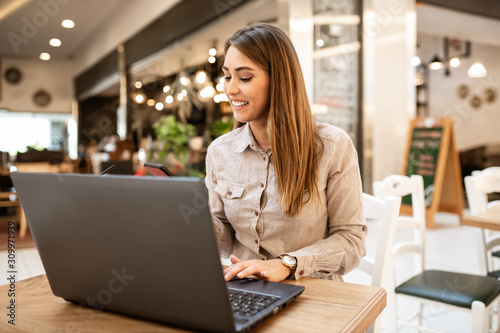 Gorgeous young caucasian woman with brown hair looking at her smart phone and smiling. There is a laptop on the table in front of her. She is sitting inside of a coffee shop.