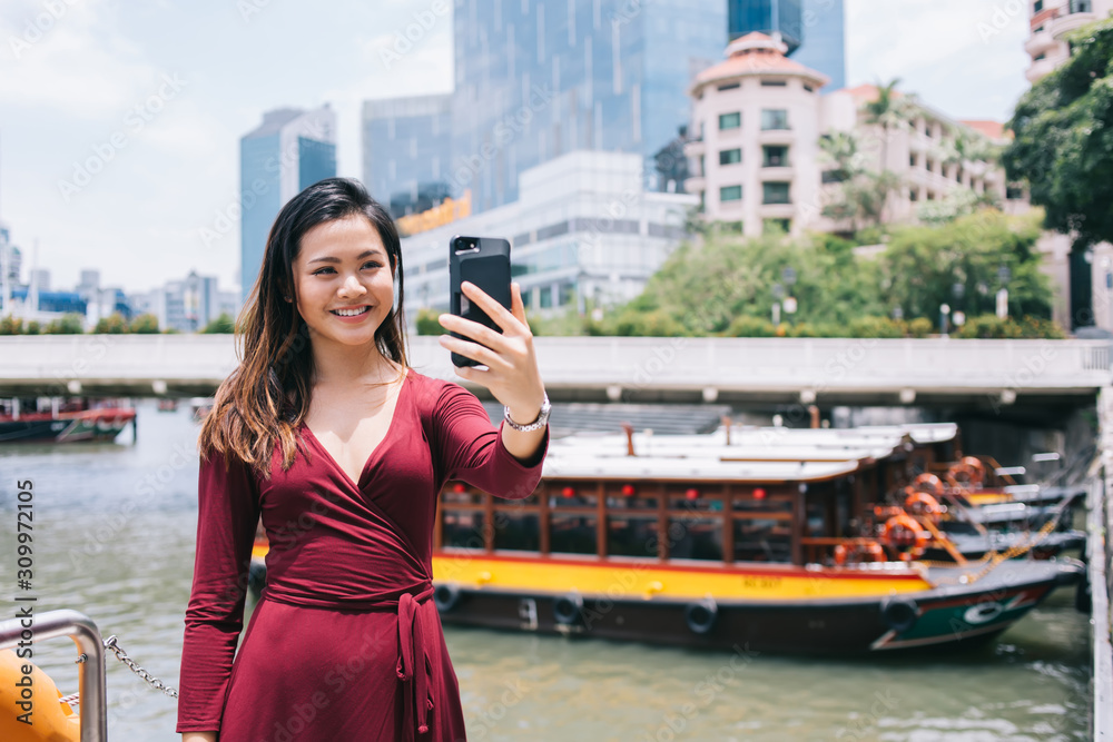 Fototapeta premium Smiling adult female capturing selfie photo by cellphone against marina