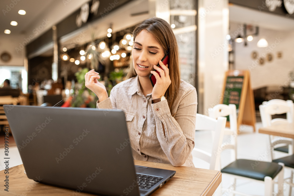 Pretty young caucasian woman with brown hair talking on her smart-phone, focused on her conversation. She is sitting in cafe. There is a laptop on the table in front of her. Pretty young caucasian woman with brown hair talking on her smart-phone, focused on her conversation. She is sitting in cafe. There is a laptop on the table in front of her.