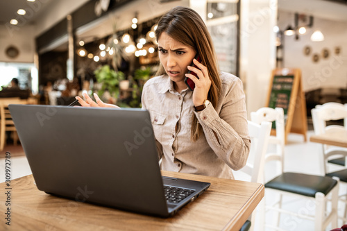Frustrated young caucasian businesswoman with brown hair talking and arguing on smart-phone with her colleague. She is angry and looking at her laptop. She is sitting in a coffee shop.