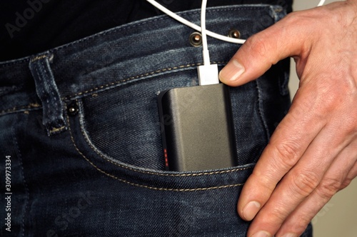 One hand putting external battery pack in jeans pocket. Power bank with led on connected to white cord in jeans pocket. Portable technology juice.