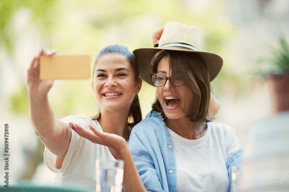 Obraz premium Two beautiful caucasian girlfriends sitting in cafe outdoors and taking selfie.