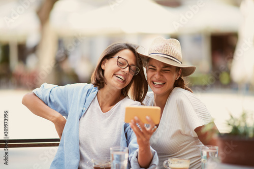 Two beautiful caucasian female friends sitting in cafe, looking at smartphone and laughing.