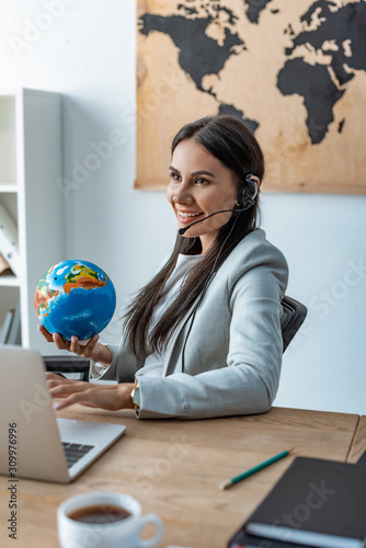 smiling travel agent holding globe while sitting at workplace photo