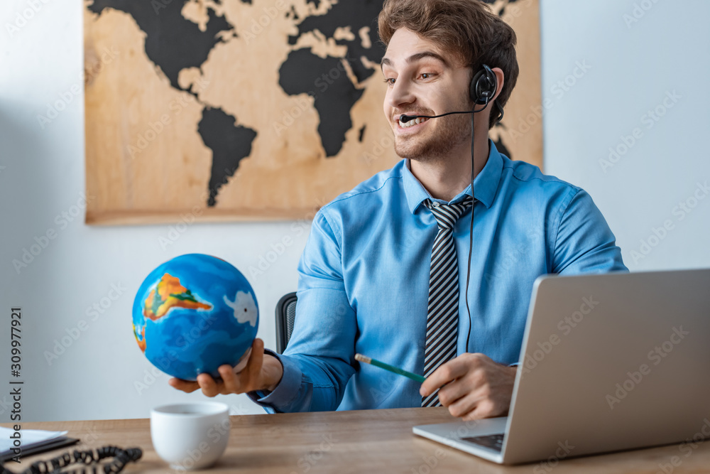smiling travel agent holding globe while sitting at workplace smiling travel agent holding globe while sitting at workplace
