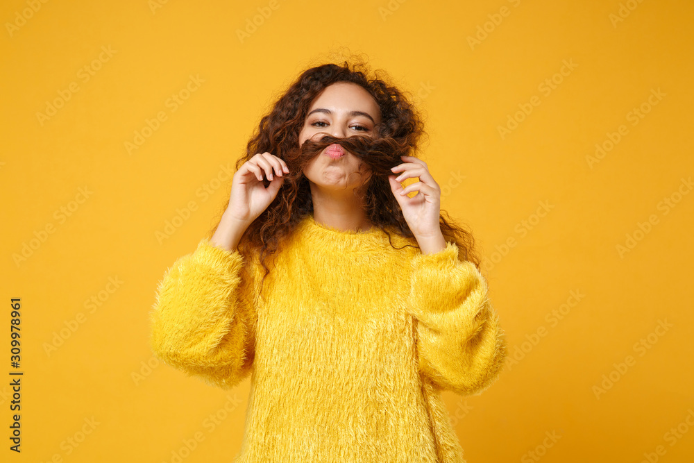 Funny young african american girl in fur sweater posing isolated on yellow orange background studio portrait. People sincere emotions lifestyle concept. Mock up copy space. Keeping hair like mustache. Funny young african american girl in fur sweater posing isolated on yellow orange background studio portrait. People sincere emotions lifestyle concept. Mock up copy space. Keeping hair like mustache.