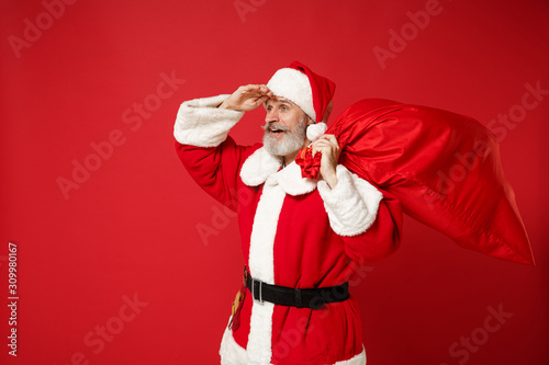 Elderly gray-haired mustache bearded Santa man in Christmas hat posing isolated on red background. New Year 2020 celebration holiday concept. Mock up copy space. Hold bag with gifts, looking far away.