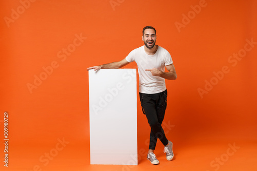 Cheerful young man in casual white t-shirt posing isolated on orange wall background in studio. People lifestyle concept. Mock up copy space. Pointing index finger on big white empty blank billboard. photo