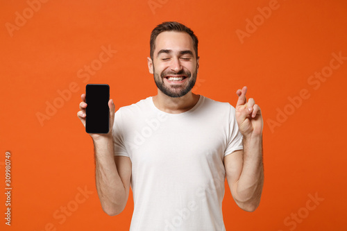 Happy man in white t-shirt posing isolated on orange background. People lifestyle concept. Mock up copy space. Holding mobile phone with blank screen keeping fingers crossed, eyes closed, making wish.