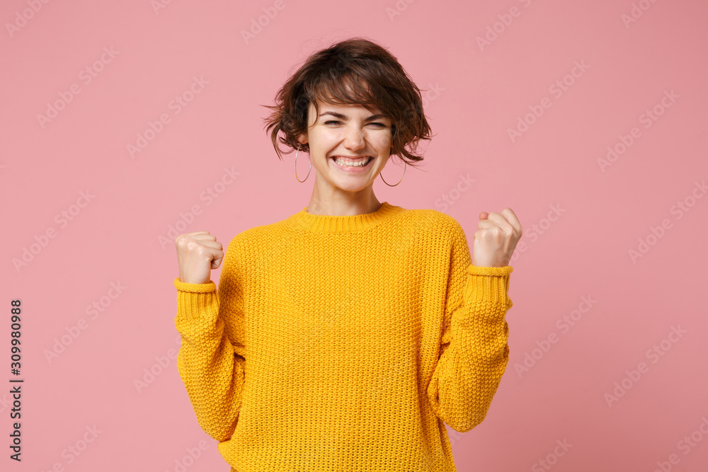 Joyful young brunette woman girl in yellow sweater posing isolated on pastel pink wall background studio portrait. People sincere emotions lifestyle concept. Mock up copy space. Doing winner gesture.