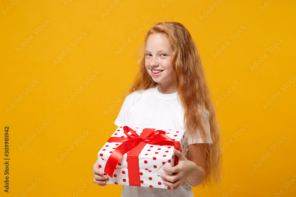 Smiling little ginger kid girl 12-13 years old in white t-shirt isolated on yellow background. New Year Women's Day birthday holiday concept. Mock up copy space. Hold present box with gift ribbon bow. Smiling little ginger kid girl 12-13 years old in white t-shirt isolated on yellow background. New Year Women's Day birthday holiday concept. Mock up copy space. Hold present box with gift ribbon bow.