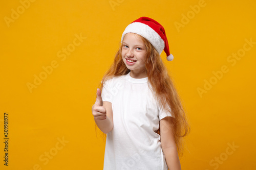 Cute little ginger kid Santa girl 12-13 years old in white t-shirt, Christmas hat isolated on yellow background. Happy New Year 2020 celebration holiday concept. Mock up copy space. Showing thumb up. © ViDi Studio
