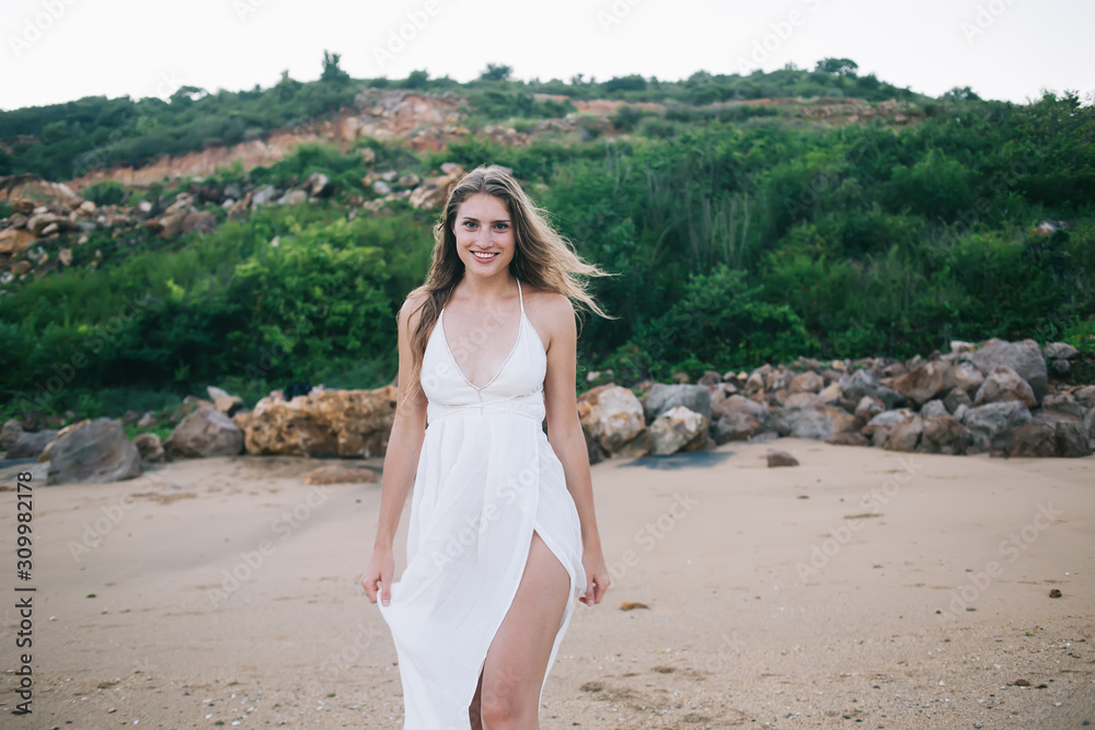 Pretty positive woman in white dress walking along beach