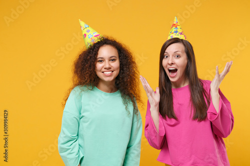 Two excited smiling women friend european african american girl in pink green clothes birthday hats posing isolated on yellow background. People lifestyle concept. Mock up copy space. Spreading hands.