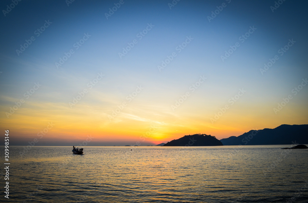 Silhouette photo of seascape with beautiful color of sunset sky, mountains, reflection of the sun on sea water and boat that going home before sky getting dark.