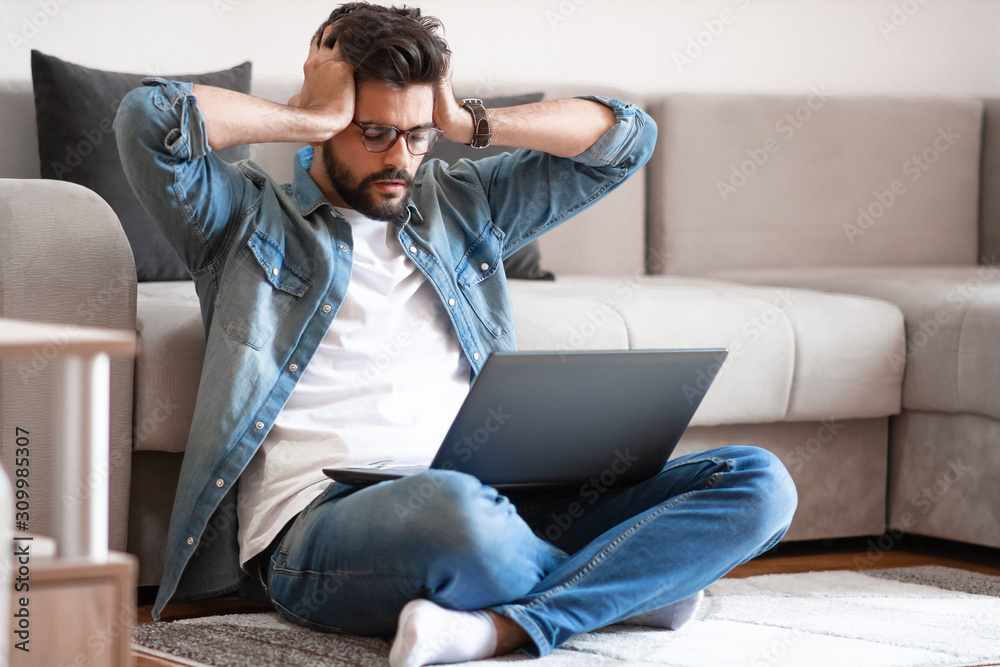Annoyed caucasian young bearded man with eyeglasses and dressed casual sitting on floor with laptop in lap, holding head and getting nervous about situation on his bank account. Annoyed caucasian young bearded man with eyeglasses and dressed casual sitting on floor with laptop in lap, holding head and getting nervous about situation on his bank account.