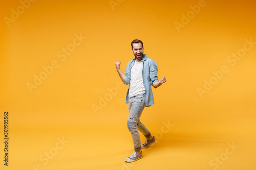 Happy young bearded man in casual blue shirt posing isolated on yellow orange background studio portrait. People sincere emotions lifestyle concept. Mock up copy space. Clenching fists like winner.