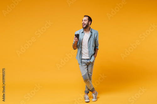 Laughing young man in casual blue shirt posing isolated on yellow orange background, studio portrait. People emotions lifestyle concept. Mock up copy space. Hold cup of coffee or tea, looking aside.