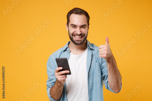 Smiling young man in casual blue shirt posing isolated on yellow orange wall background in studio. People lifestyle concept. Mock up copy space. Using mobile phone typing sms message showing thumb up.