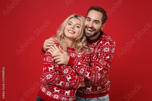 Cheerful young couple guy girl in Christmas knitted sweaters posing isolated on red wall background studio portrait. Happy New Year 2020 celebration holiday party concept. Mock up copy space. Hugging.