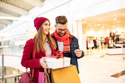 Cheerful young caucasian woman holding Christmas gift which her boyfriend gave her. She's carrying shopping bags while they are standing in a shopping mall.