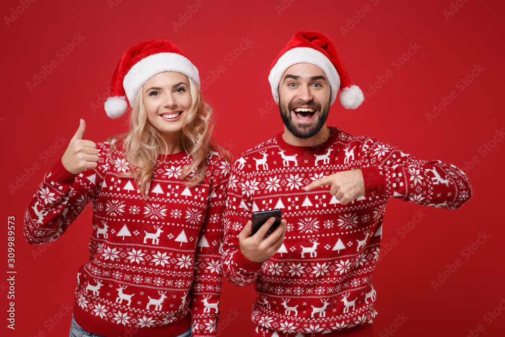 Young couple guy girl in Christmas sweaters, Santa hat posing isolated on red background. New Year 2020 celebration party concept. Mock up copy space. Pointing finger on mobile phone showing thumb up. Young couple guy girl in Christmas sweaters, Santa hat posing isolated on red background. New Year 2020 celebration party concept. Mock up copy space. Pointing finger on mobile phone showing thumb up.