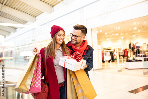 Cheerful good-looking hipster boyfriend giving his attractive surprised girlfriend a Christmas gift. They are standing in a shopping mall while she's carrying shopping bags.