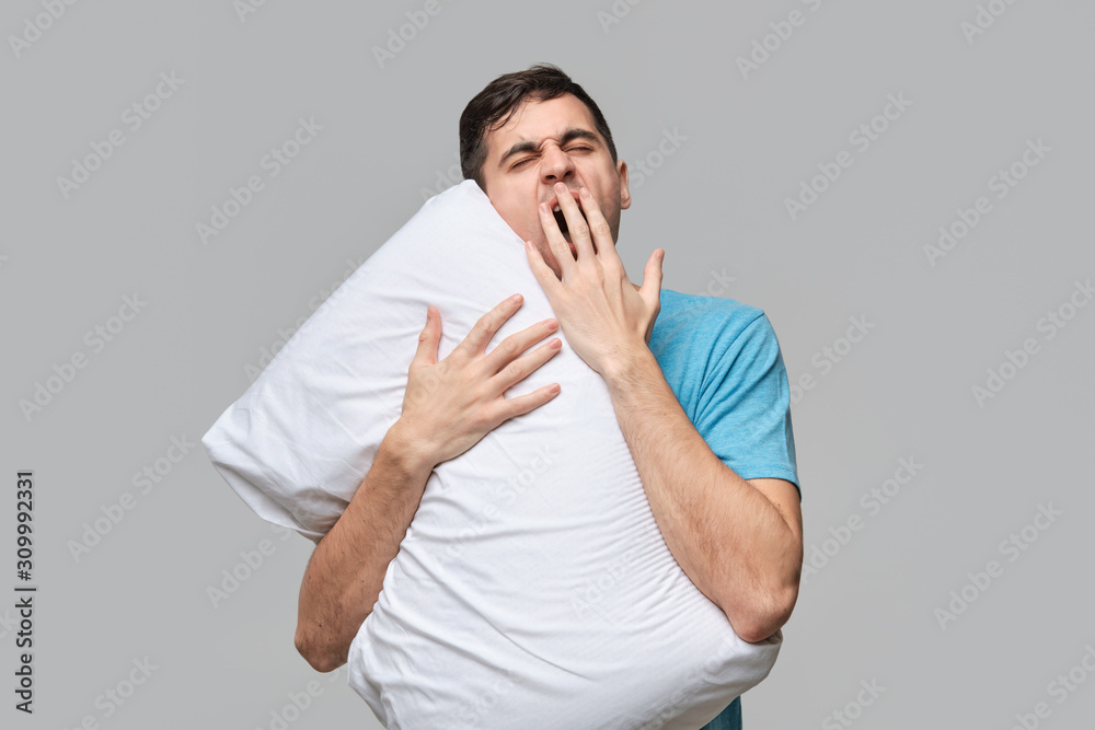 Tired brunet man in a blue tee yawning hugging white pillow isolated over grey background. Tired brunet man in a blue tee yawning hugging white pillow isolated over grey background.