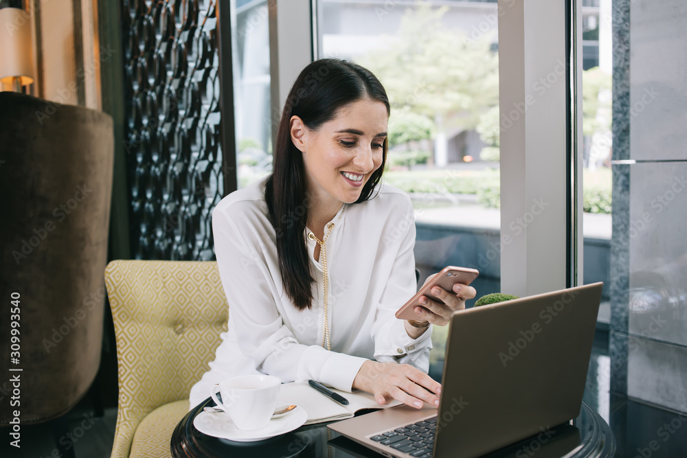 Woman browsing smartphone with smile at table with laptop in cafe Woman browsing smartphone with smile at table with laptop in cafe