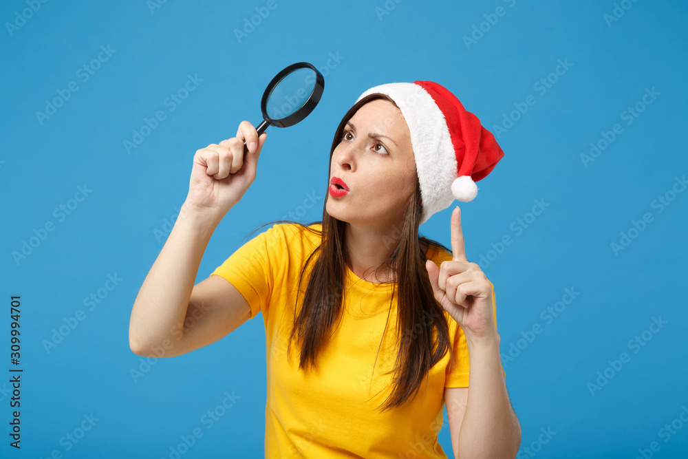 Serious young woman Santa girl in Christmas hat posing isolated on blue background. Happy New Year 2020 celebration holiday concept. Mock up copy space. Hold magnifying glass pointing index finger up. Serious young woman Santa girl in Christmas hat posing isolated on blue background. Happy New Year 2020 celebration holiday concept. Mock up copy space. Hold magnifying glass pointing index finger up.