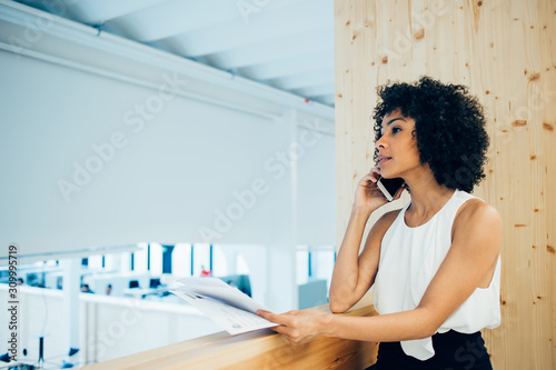 Fototapeta Naklejka Na Ścianę i Meble -  Dark skinned female proud ceo calling on smartphone to solve financial issues in accounting reports, attractive businesswoman with documents in hands talking on mobile phone device in office interior