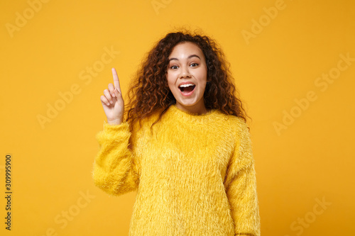 Excited young african american girl in fur sweater posing isolated on yellow orange background, studio portrait. People lifestyle concept. Mock up copy space. Hold index finger up with great new idea. © ViDi Studio