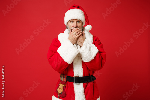Shocked elderly gray-haired mustache bearded Santa man in Christmas hat posing isolated on red background. Happy New Year 2020 celebration holiday concept. Mock up copy space. Cover mouth with hands.