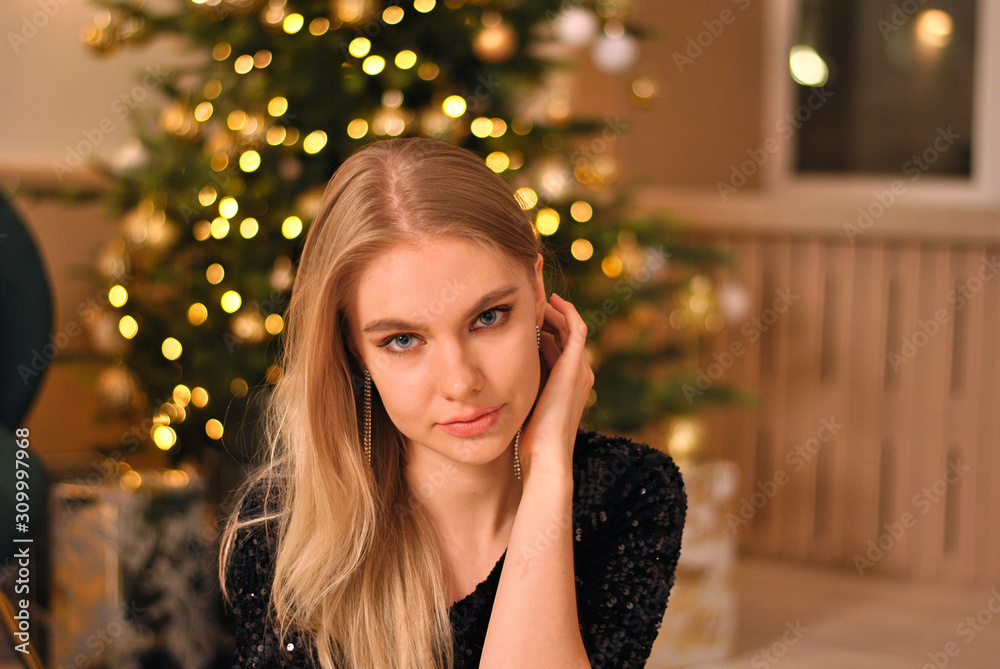 Beautiful blonde woman nearby the Christmas tree closeup portrait with garland lights in bokeh background Beautiful blonde woman nearby the Christmas tree closeup portrait with garland lights in bokeh background