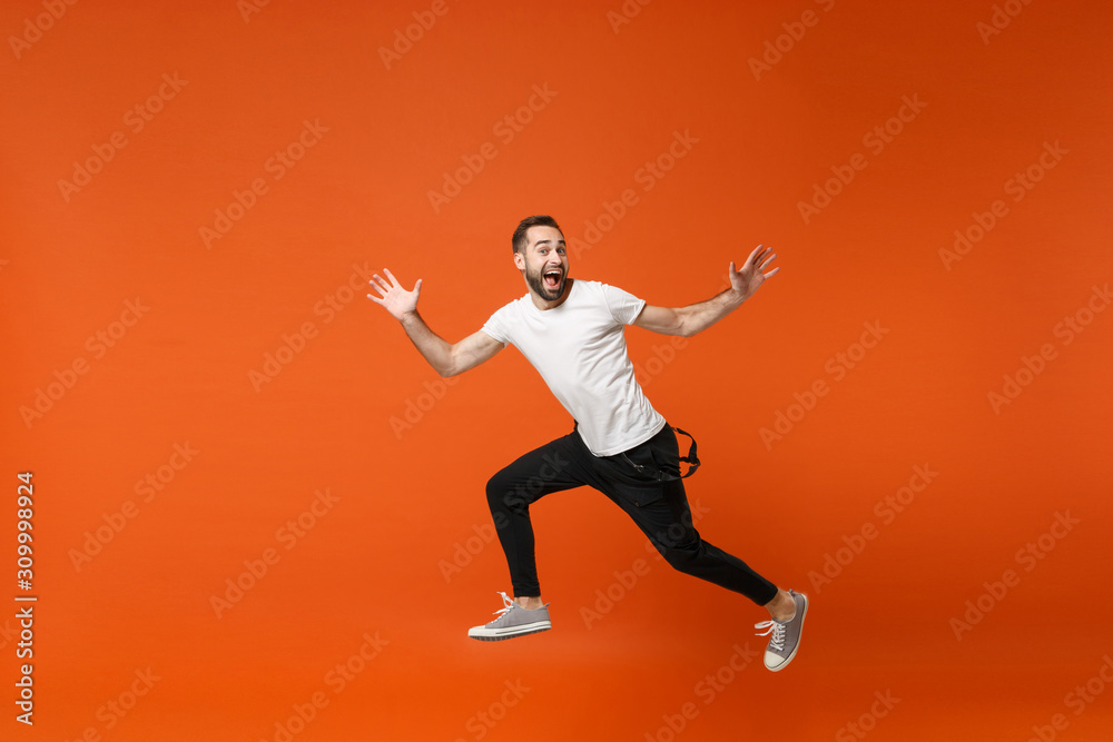 Excited young man in casual white t-shirt posing isolated on orange wall background studio portrait. People lifestyle concept. Mock up copy space. Having fun, fooling around, jumping, spreading hands.
